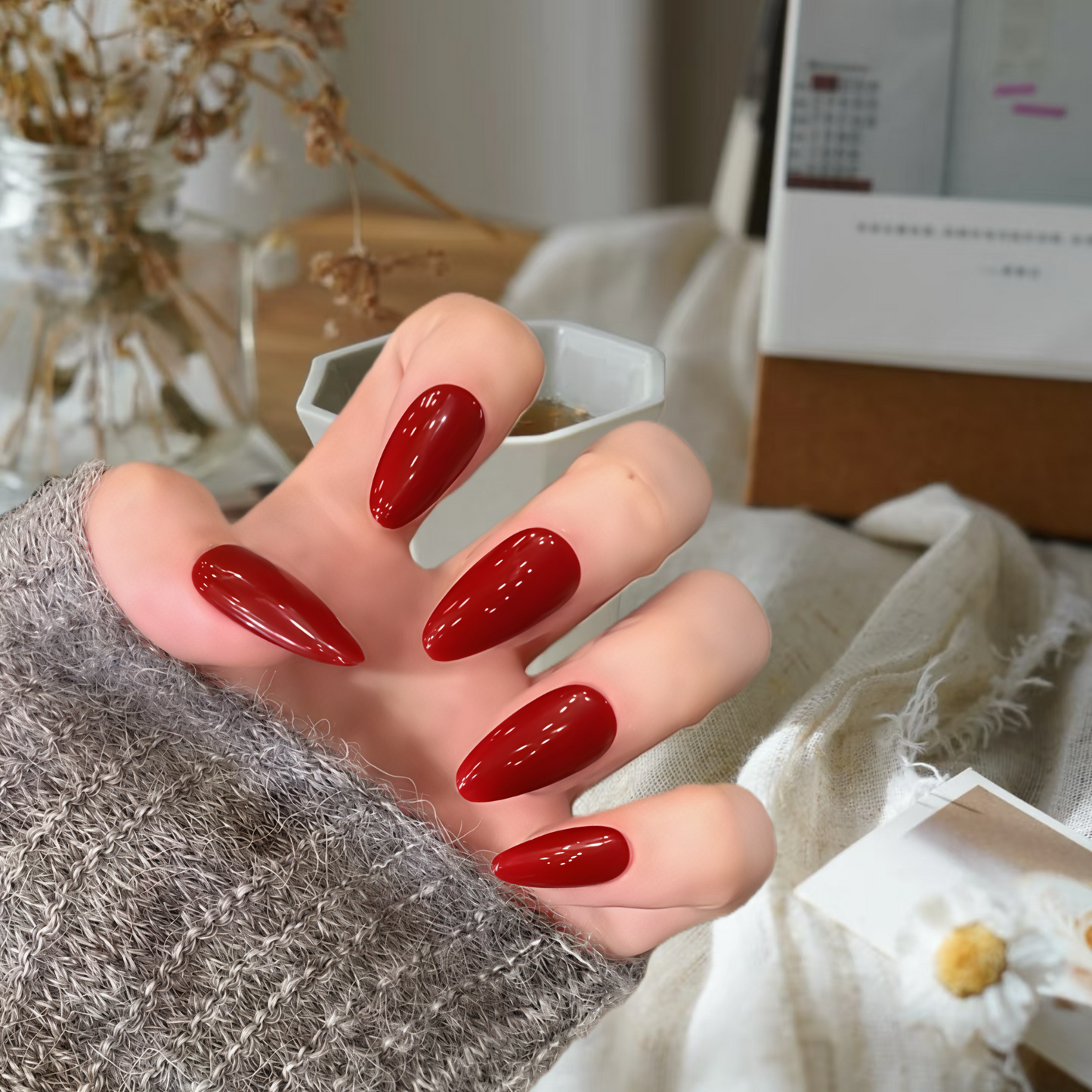 Hand with red nail polish holding a small white container, blurred indoor background