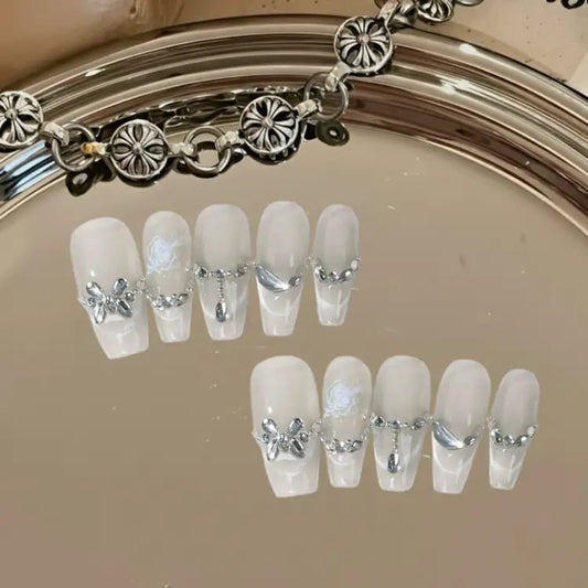 Set of decorative false nails with silver bow and chain designs on a reflective surface.