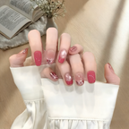 Close-up of a hand with pink and clear nail polish, wearing a white sleeve, with a blurred background.