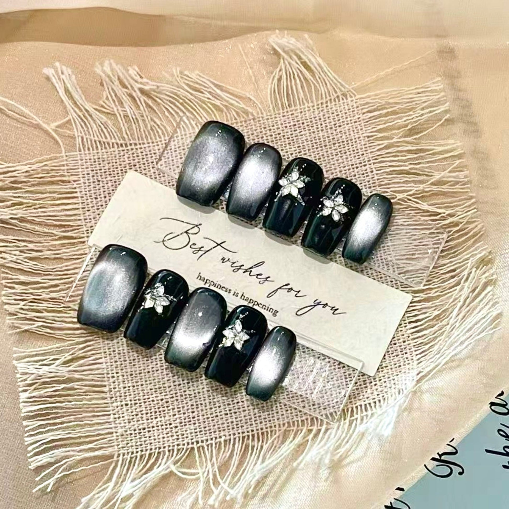 Black nail art with floral designs on a textured beige surface with a card.