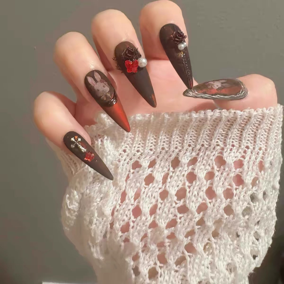 A close-up of a hand wearing long stiletto-shaped nails with a dark brown and orange gradient design, floral and bunny motifs, and a lace sleeve against a neutral gray background.