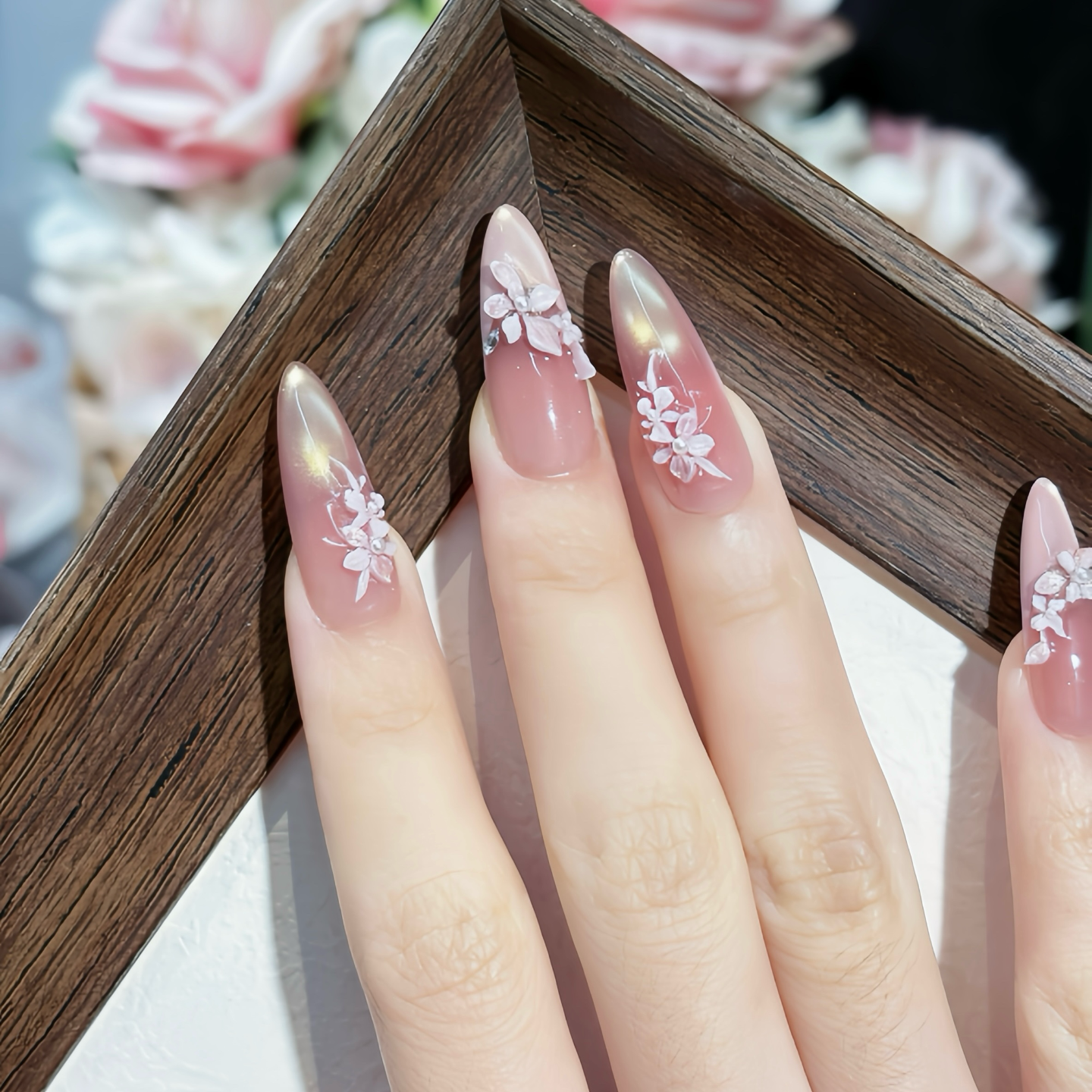 Close-up of a hand with pink nail polish and floral designs, blurred flowers in the background