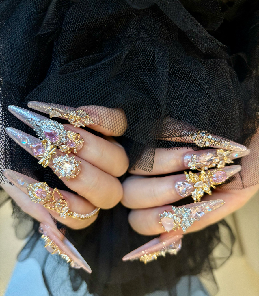 Close-up of hands with long, decorative nails and multiple rings against a black mesh background.
