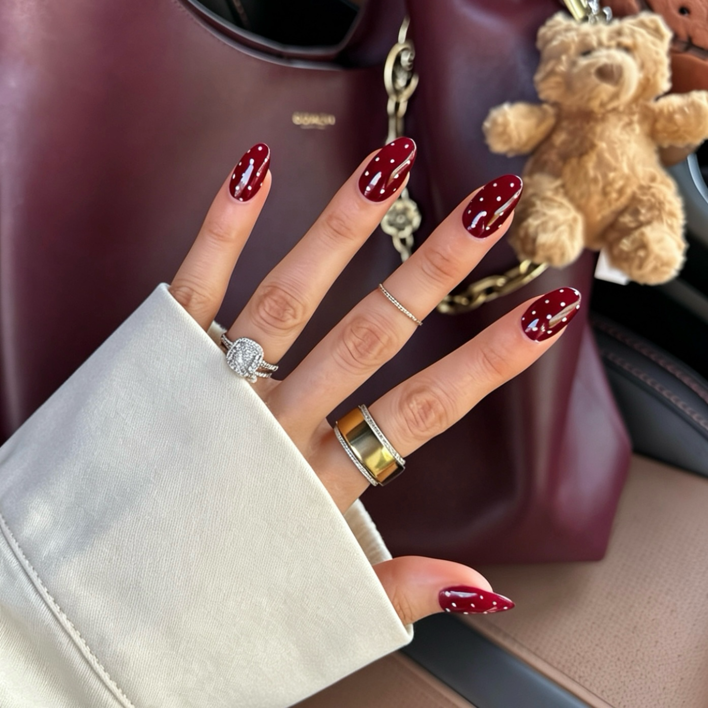 Hand with red-polka dot nails holding a white box, with a burgundy handbag and teddy bear keychain in the background.