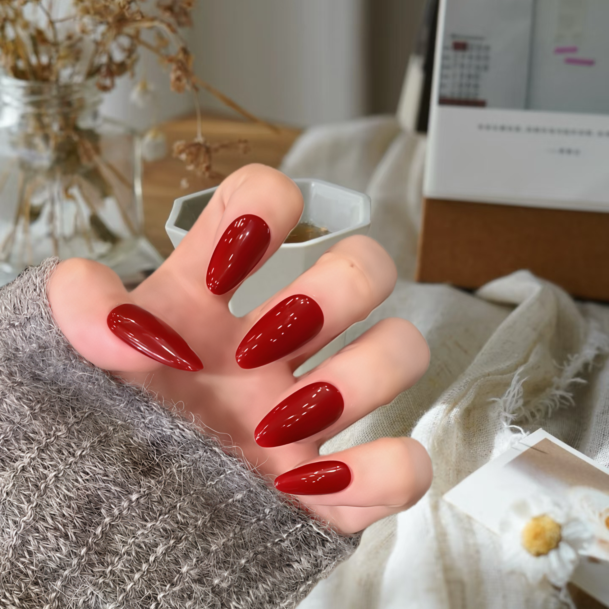 Hand with red nail polish holding a small white container, blurred indoor background