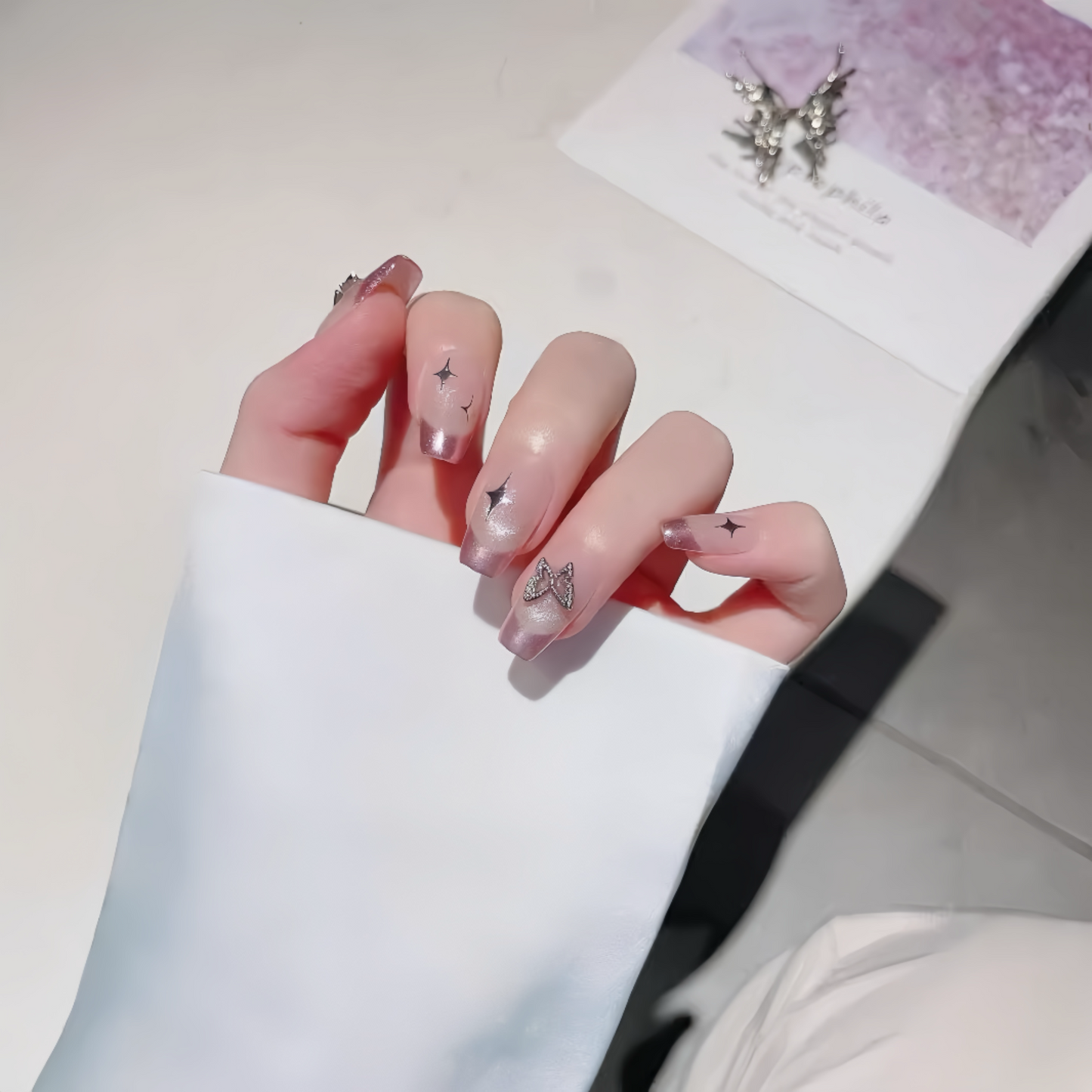 Hand with pink nail polish and butterfly designs, wearing a white sleeve, on a light background.