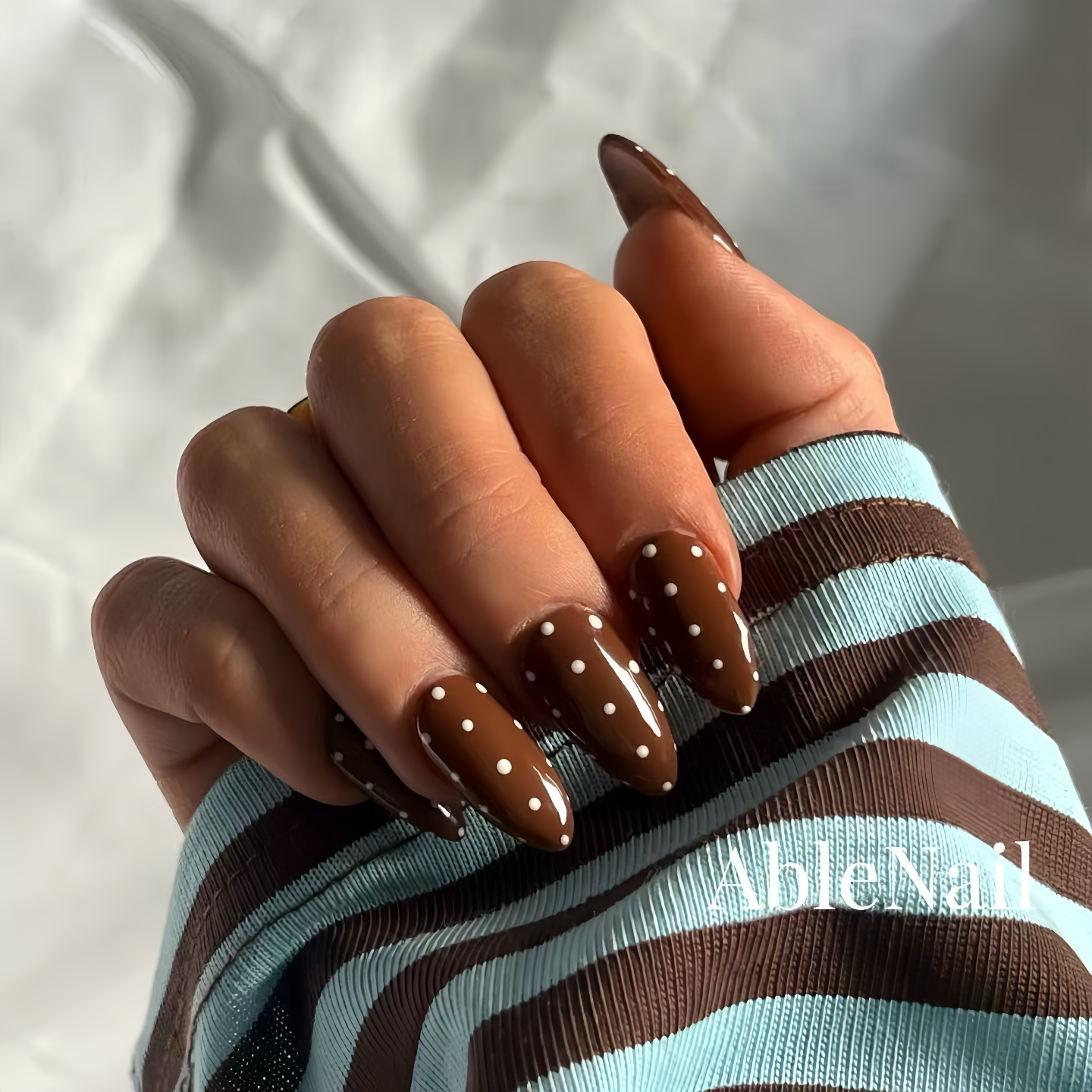 Hand with brown nail polish and polka dots, wearing a striped sleeve, on a neutral background.