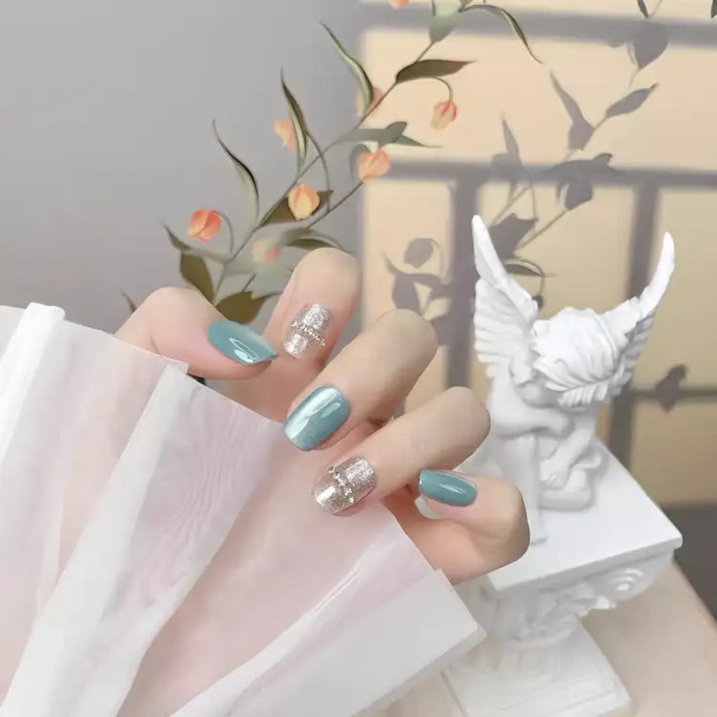 Hand with light blue and silver nail polish holding a white fabric, with a decorative background.