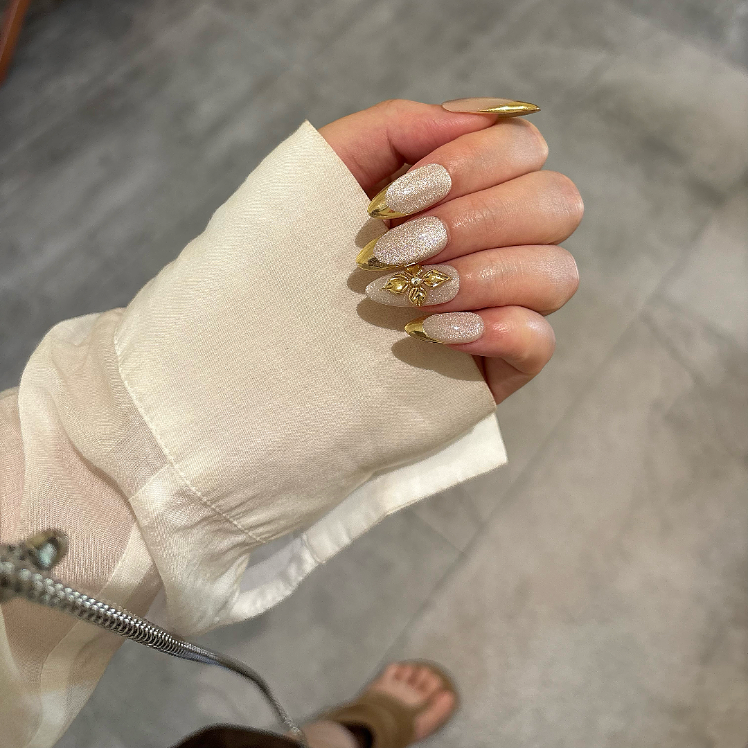 Hand with gold and white nail design wearing a beige sleeve on a wooden floor.
