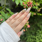 Hand with beige and gold nail design against a green leafy background