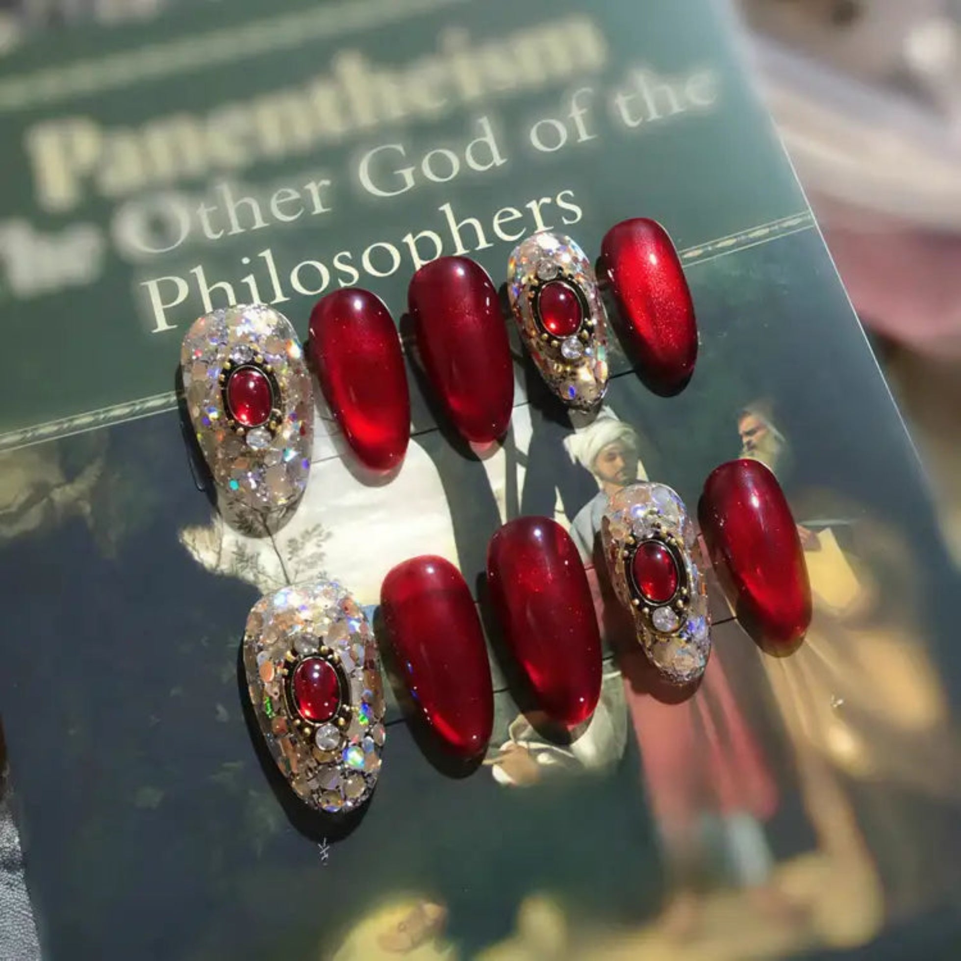 Red nail polish with decorative designs on a book titled 'Other God of the Philosophers'.