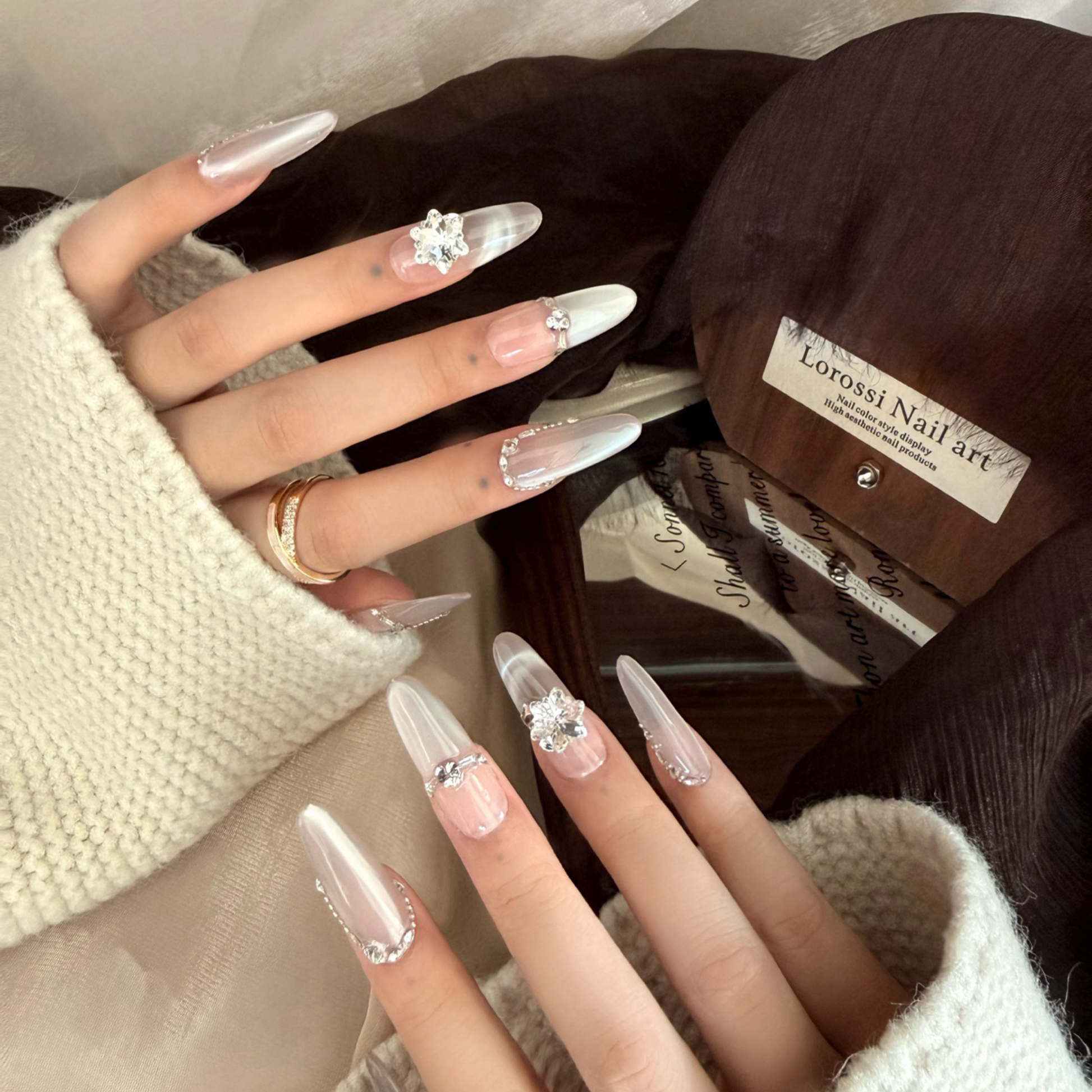 Close-up of hands with clear nail polish and rhinestone decorations, wearing a white sweater and brown gloves.