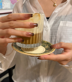 A close-up lifestyle shot of a person holding a glass of layered coffee on a saucer. The person is wearing a white button-down shirt and has long, manicured brown nails.