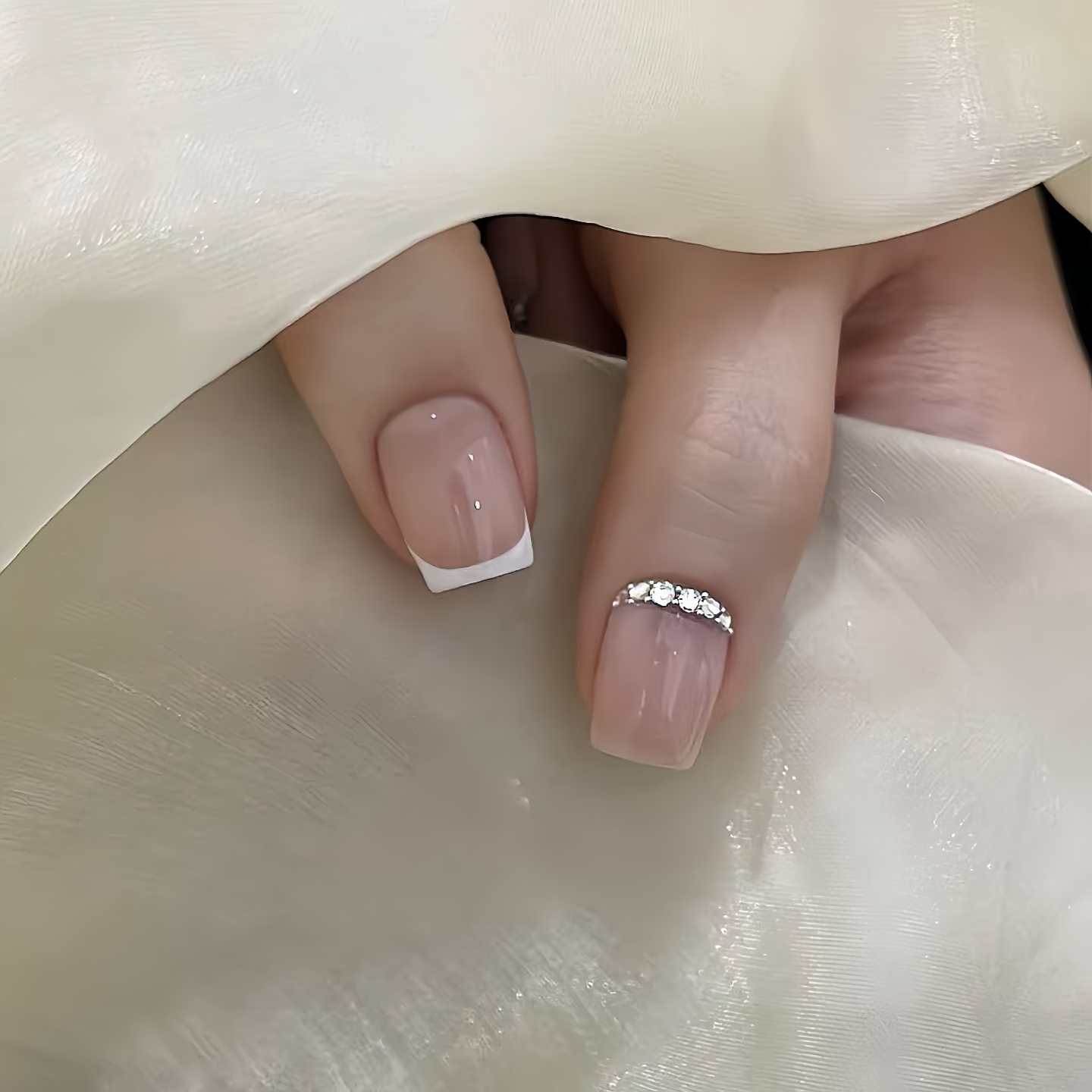 Close-up of a hand with pink nail polish and a diamond ring on a white fabric background