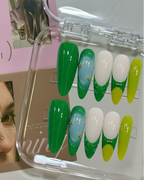 Set of green, white, and yellow false nails in a clear case with a magazine in the background.