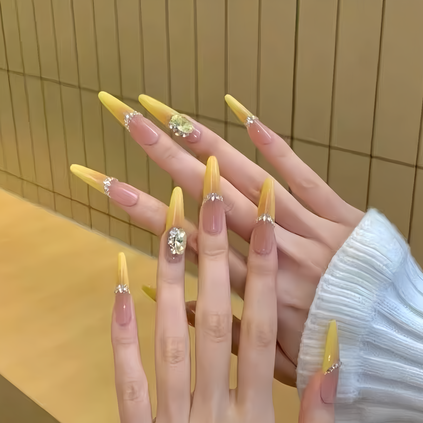 Close-up of a hand with yellow acrylic nails featuring rhinestones against a yellow tiled wall.