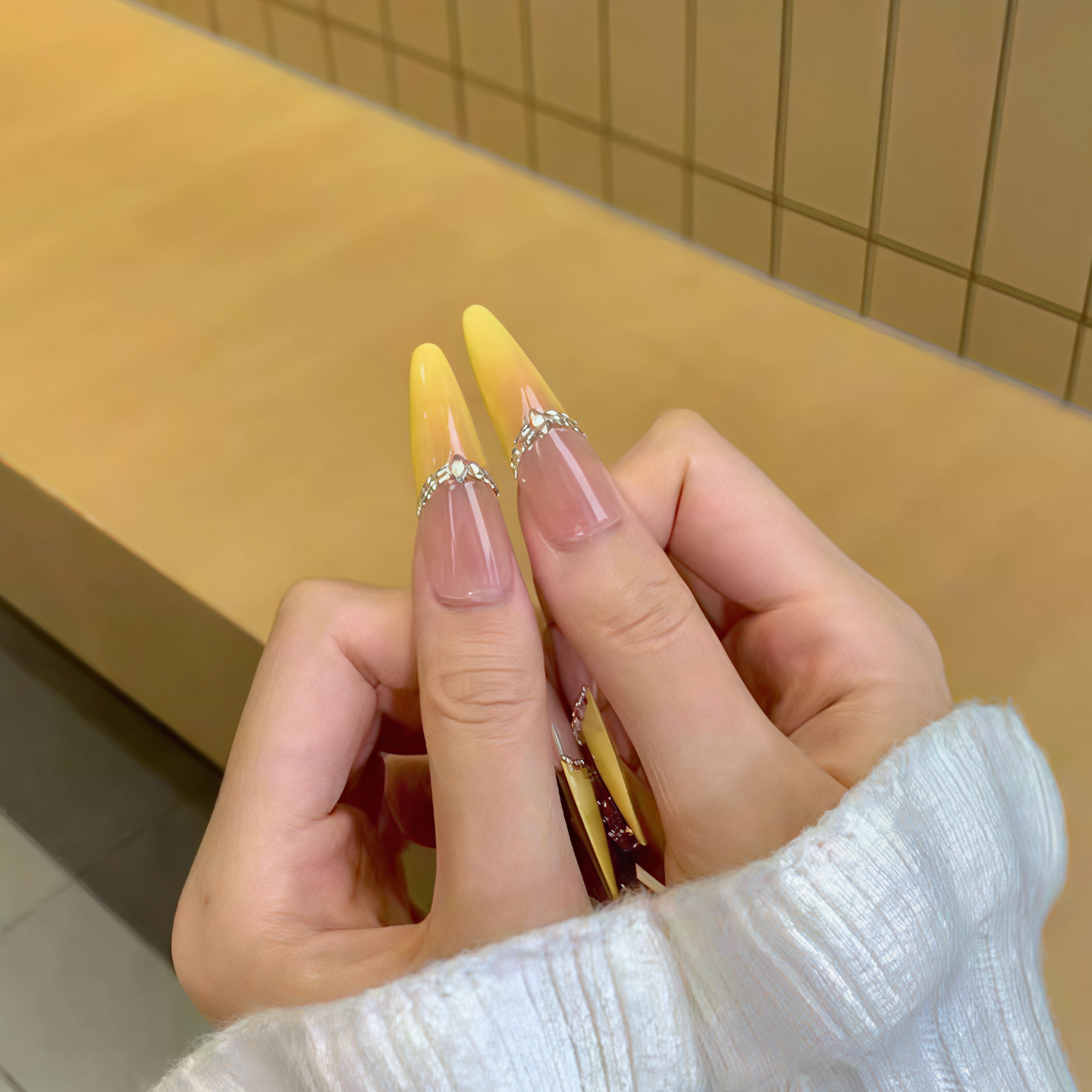 Close-up of a hand with yellow and pink nail polish holding a small object against a tiled wall background.