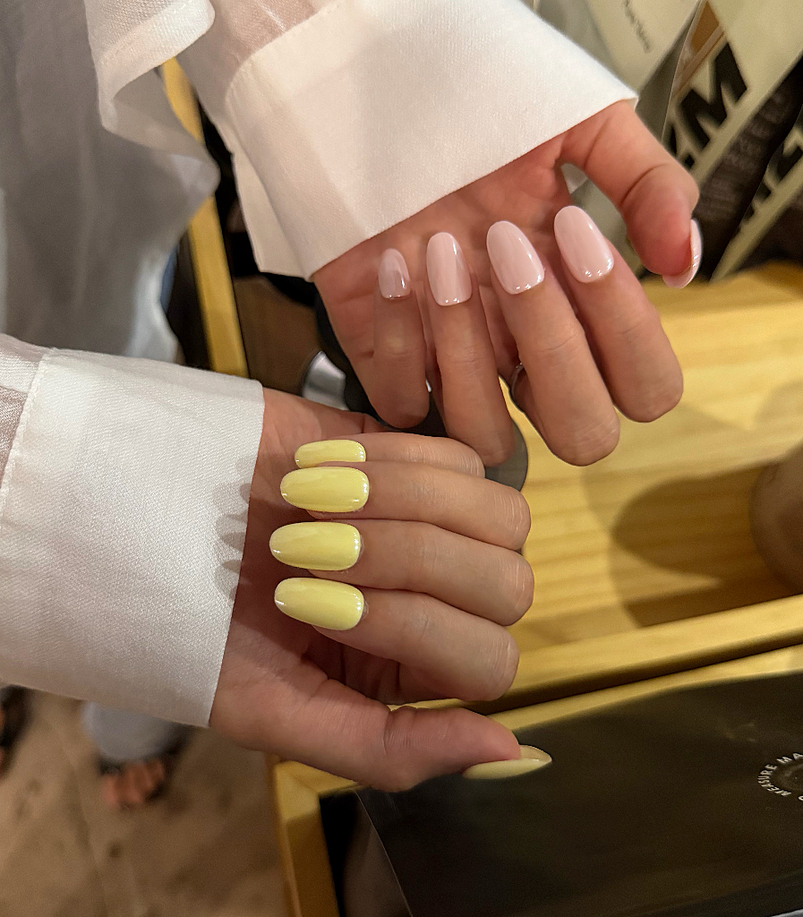 A close-up of two hands displaying manicured nails. One hand has glossy pale pink nails, and the other has glossy pale yellow almond-shaped nails.