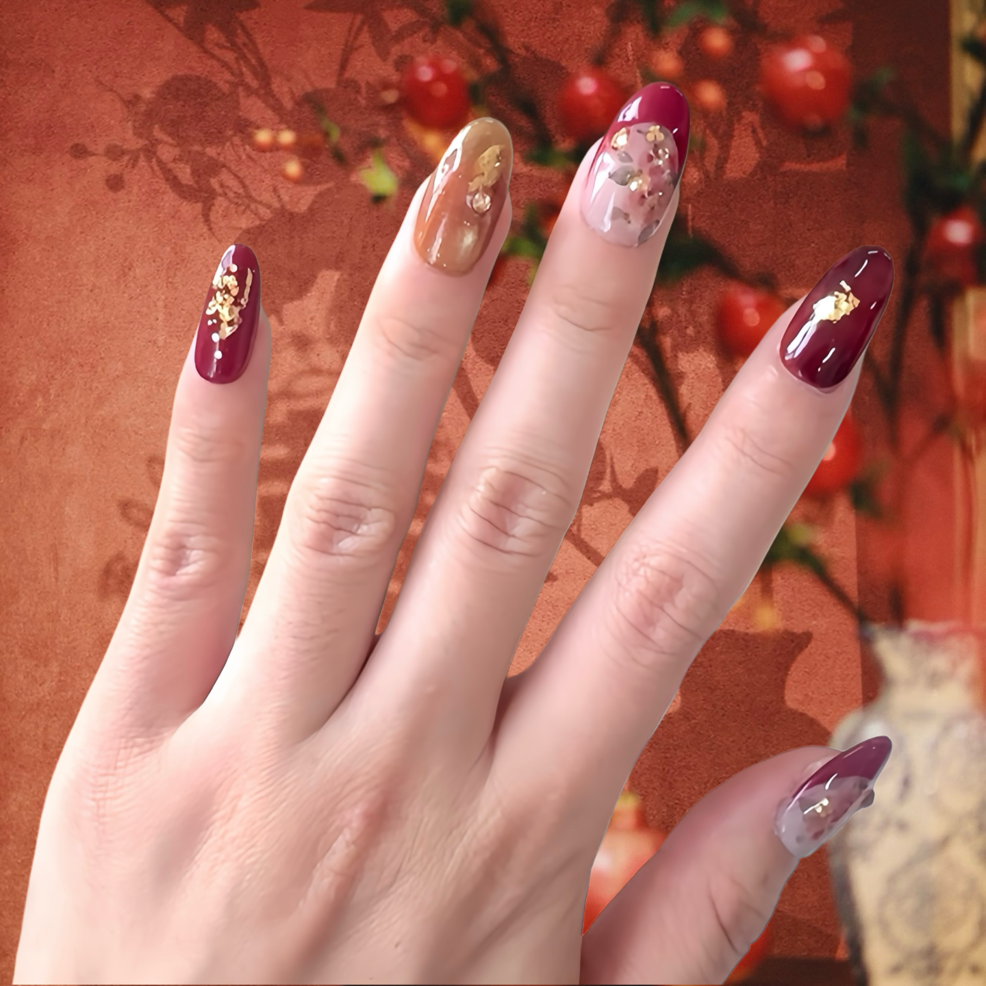 Hand with maroon nail polish and gold accents against a decorative background