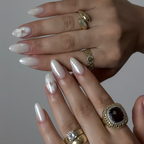 Close-up of a hand with long, manicured nails and multiple gold rings on a neutral background