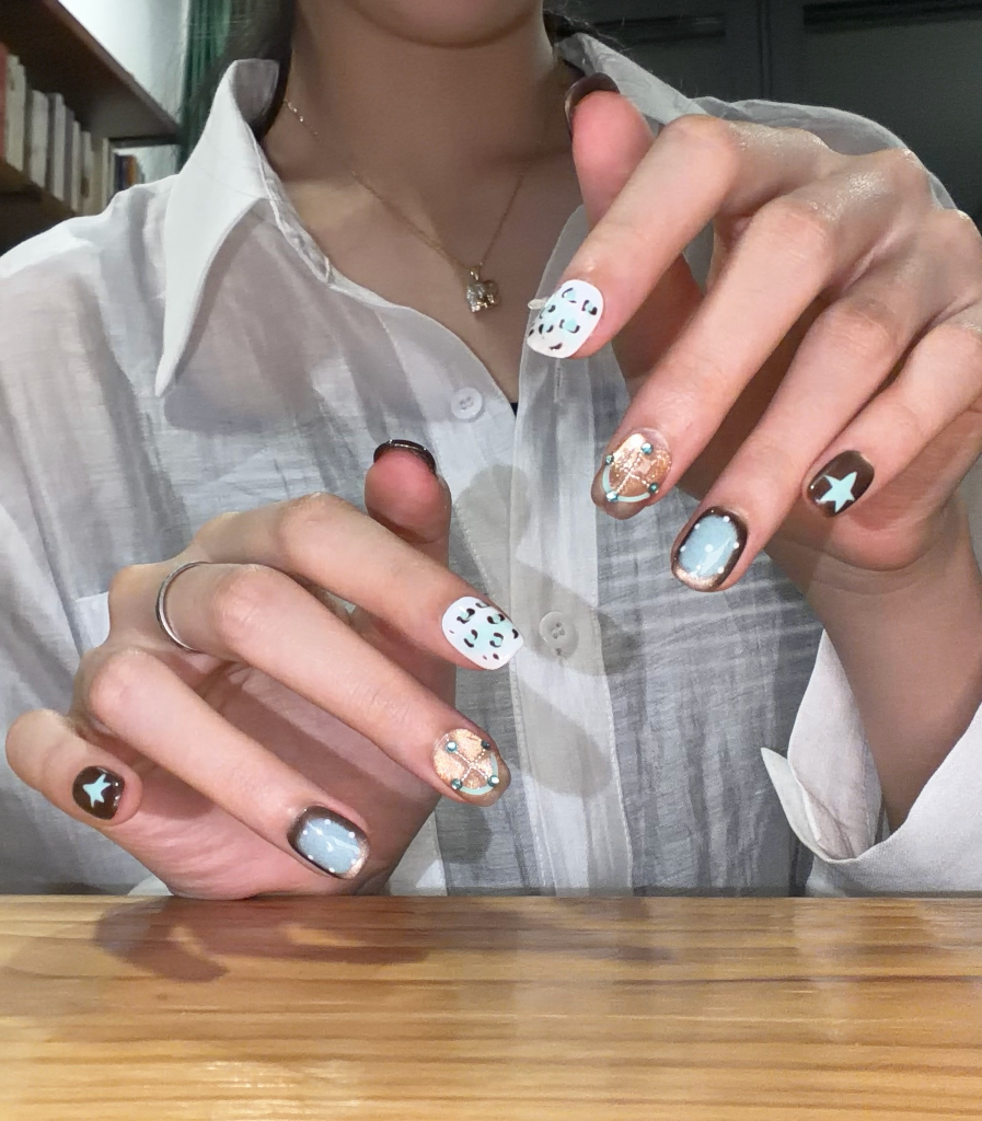 A close-up of a person's hands showcasing a detailed nail art set. The nails feature a mix of solid light blue, white with black dots, brown with gold glitter, and dark brown with a small star.