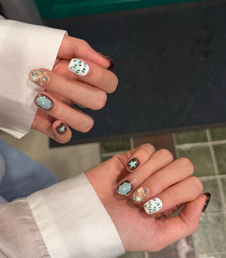 Close-up of two hands displaying a set of decorative press-on or manicured nails. The nails feature a mix of solid white, light blue-gray, dark brown, and clear glittery designs with small green leaf-like patterns.