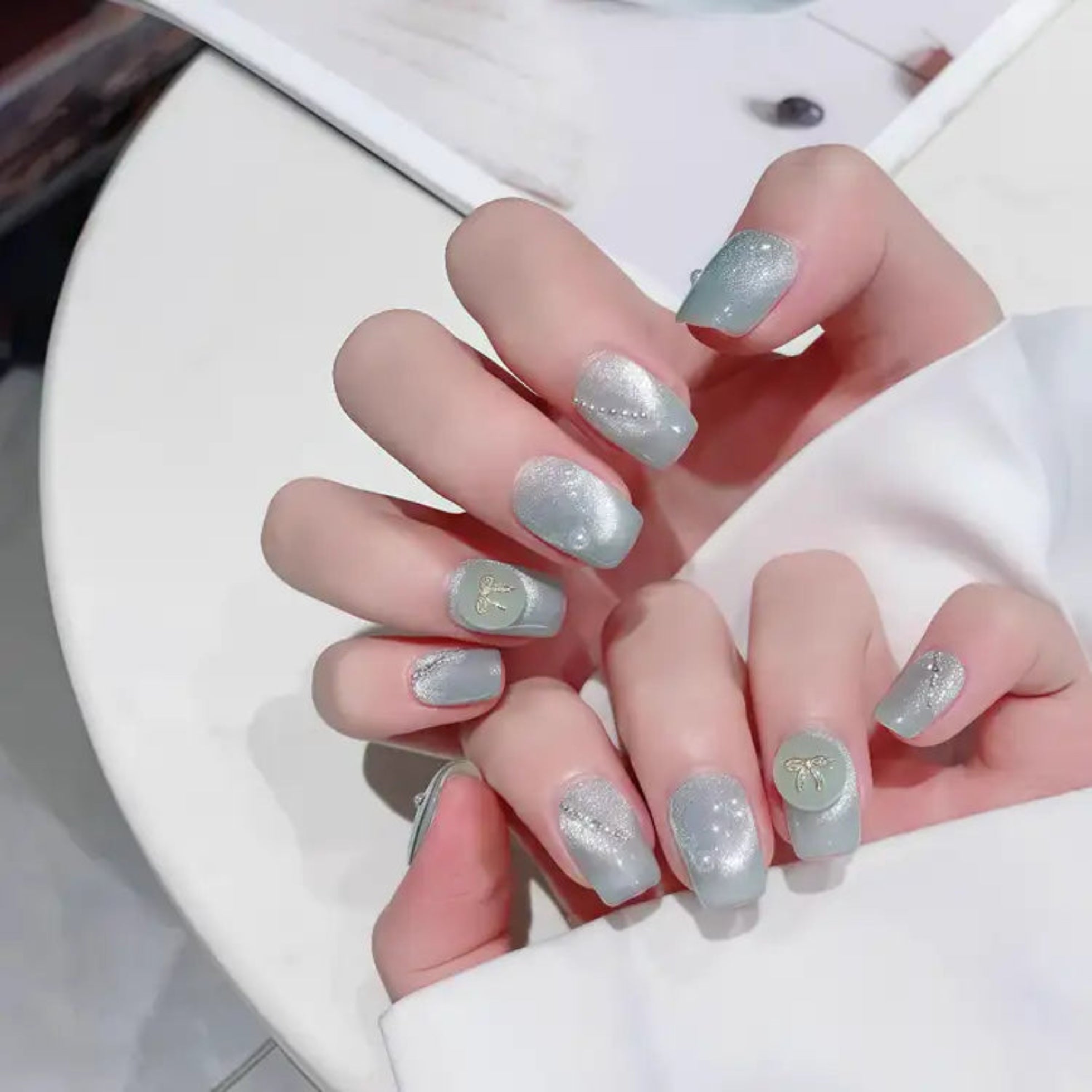 Close-up of a hand with light gray nail polish and glittery accents on a white surface.