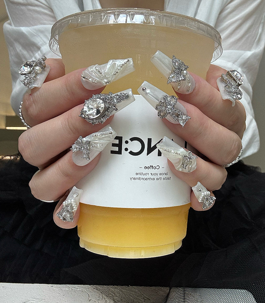 Person holding a cup with decorative nails and jewelry, wearing a white shirt.