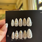 Set of white nail extensions with gold accents on a black card held by a hand.