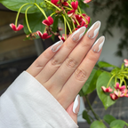 Hand with almond-shaped nails featuring a white and beige design, held against a natural background with green leaves and pink flowers.