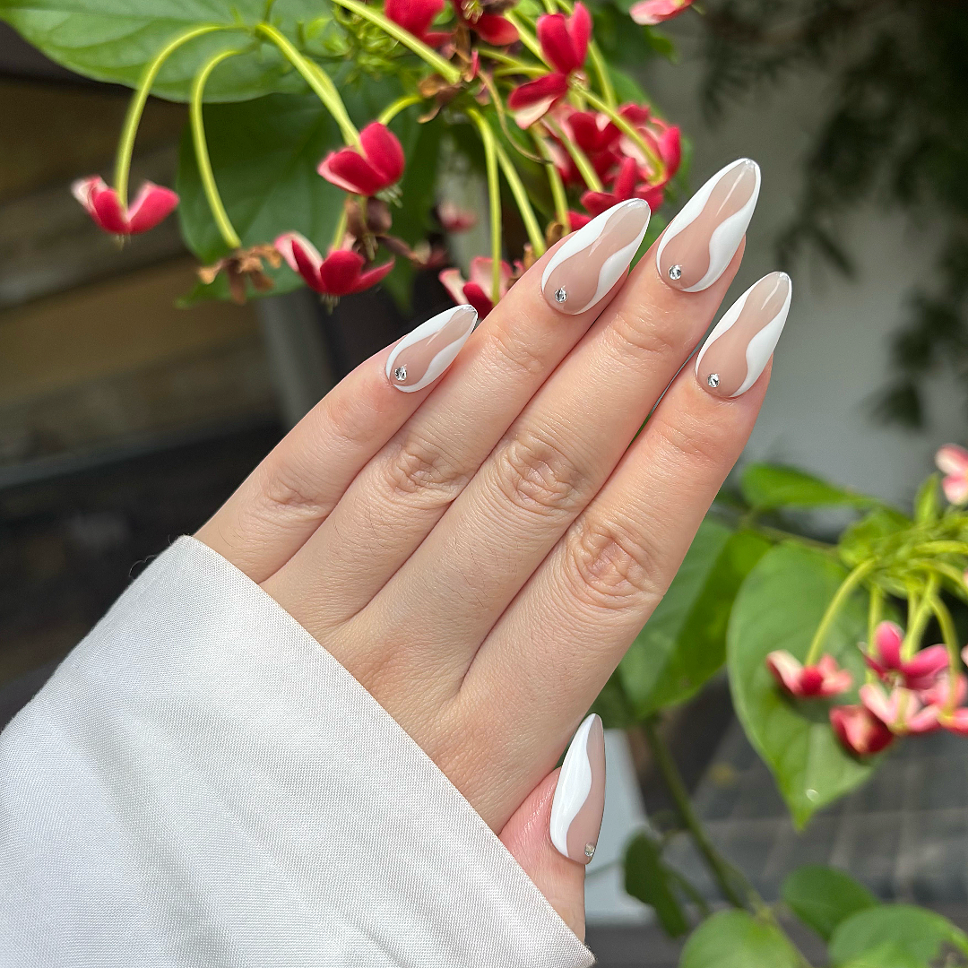 Hand with almond-shaped nails featuring a white and beige design, held against a natural background with green leaves and pink flowers.