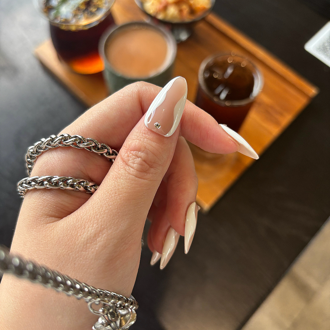 Hand with silver rings and beige nail polish holding a glass of red wine, blurred background of drinks on a tray.