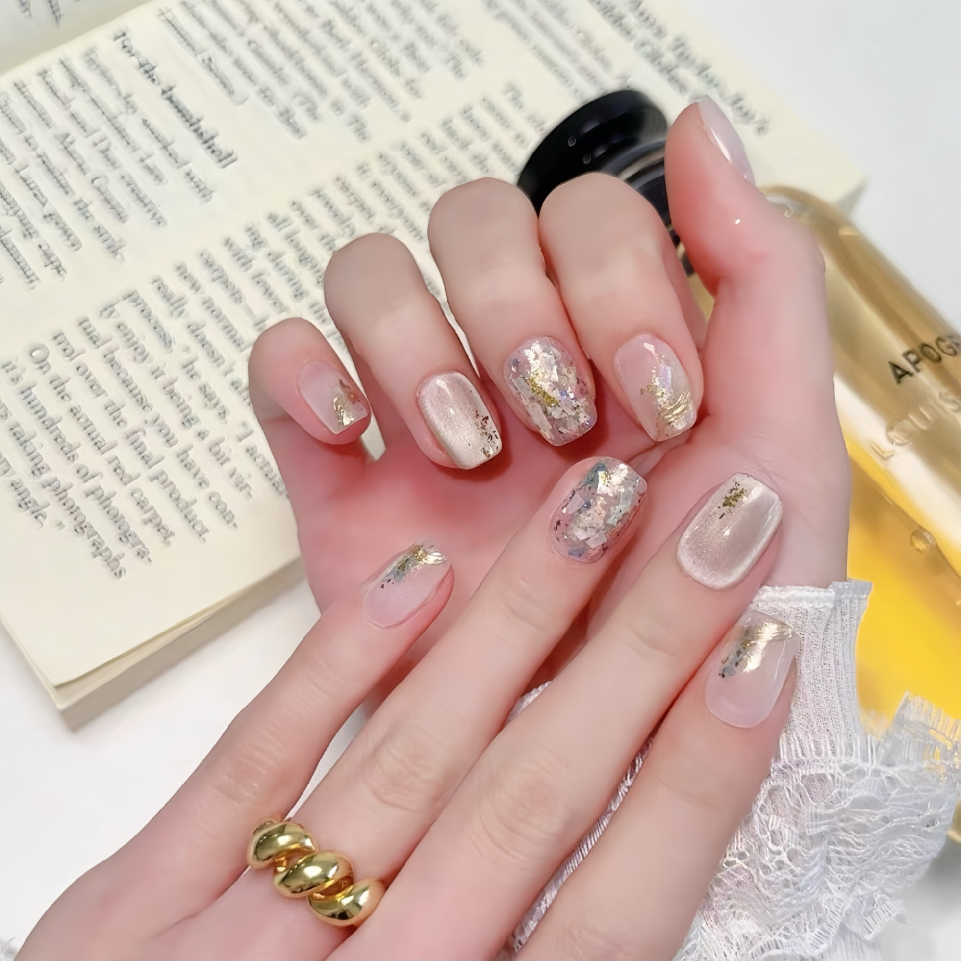 Hand with decorative nails holding a bottle of nail polish, with a book in the background.