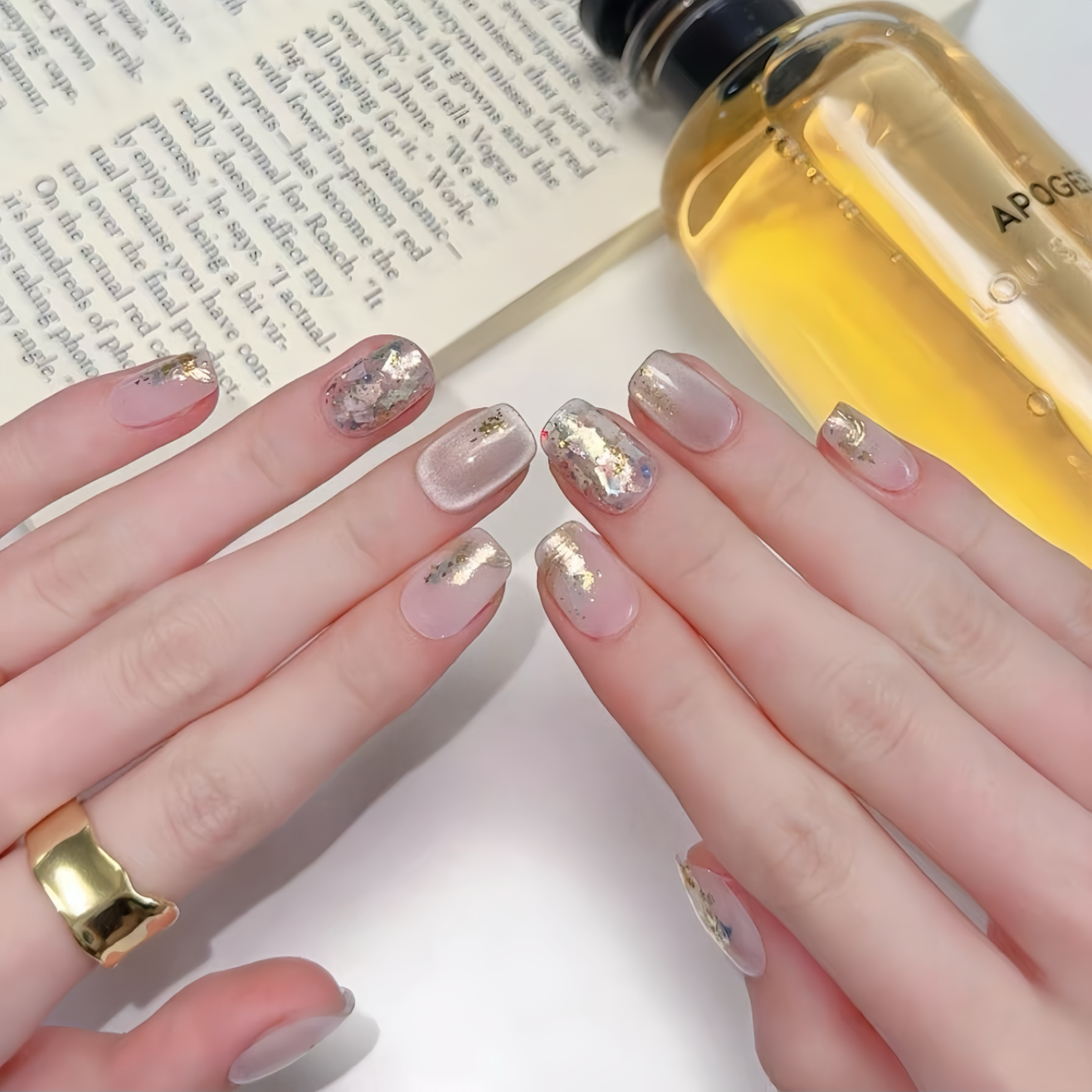 Close-up of hands with decorative nails holding a bottle of oil next to an open book.