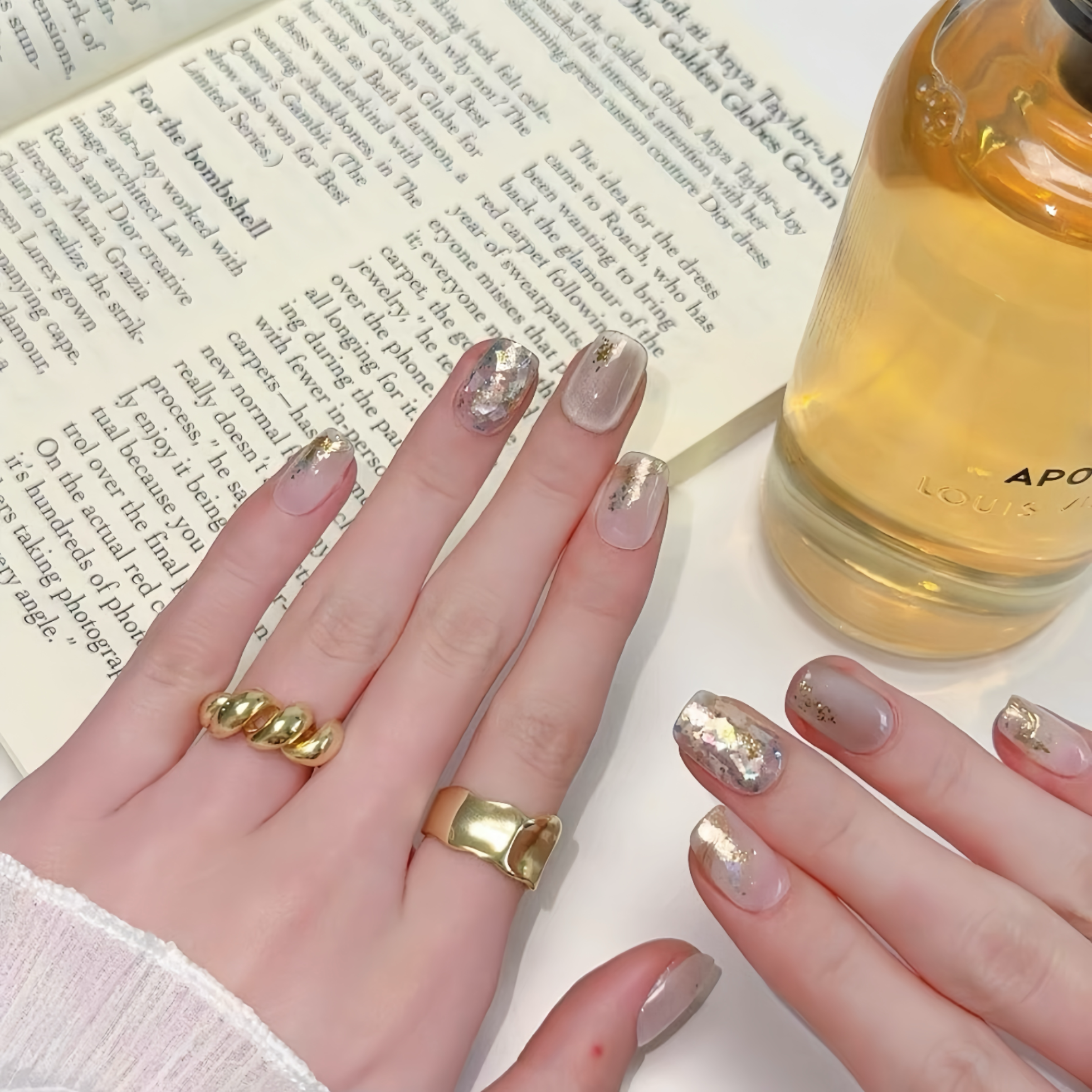 Close-up of hands with gold rings and decorative nails, next to a book and bottle.