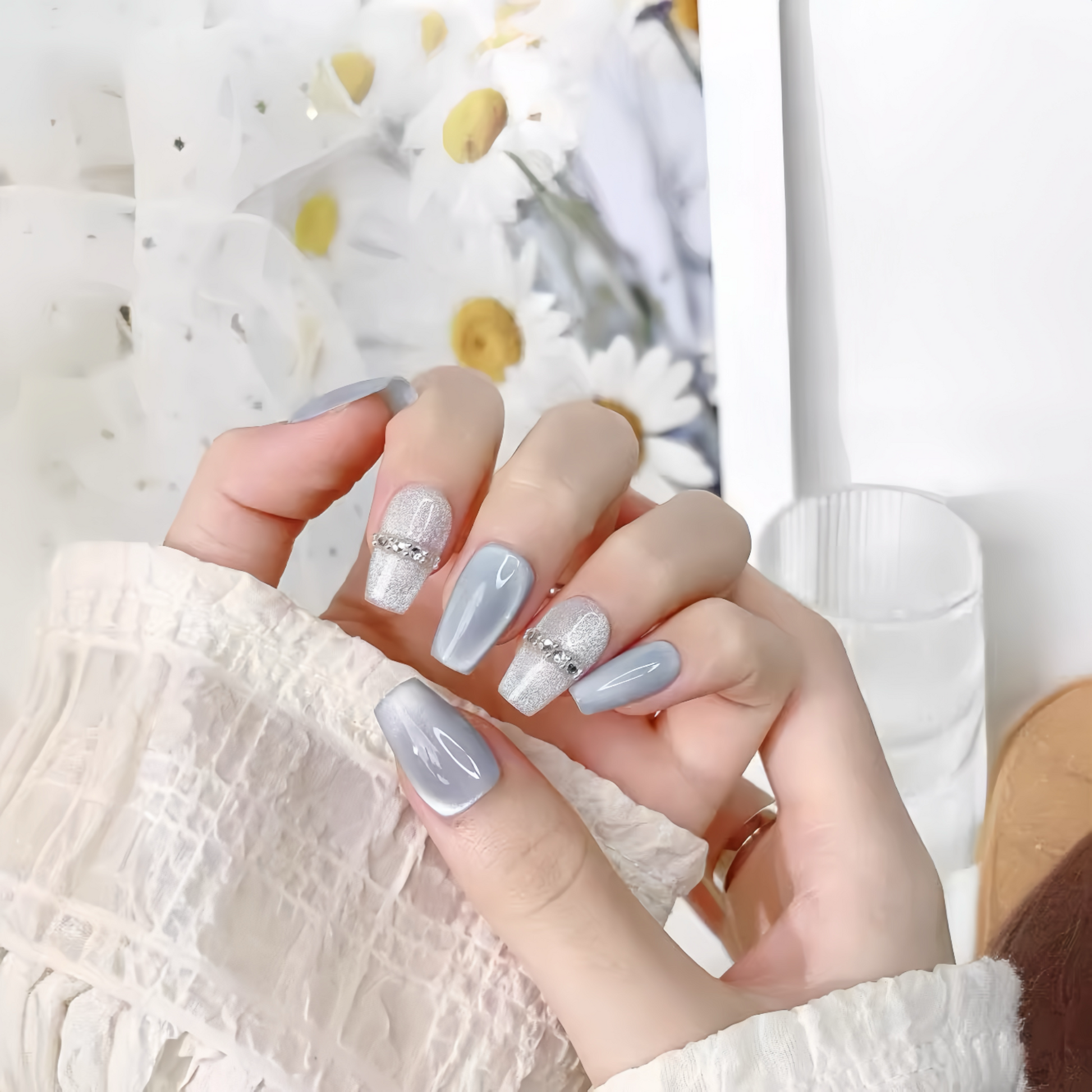 Hand with light gray nail polish and decorative rings against a blurred floral background