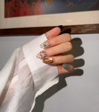 A close-up of a hand with decorative press-on nails, resting against a white sheer sleeve. The nails feature a mix of matte sage green, gold foil, white daisy designs, and a dark brown accent, set against a background with a framed picture.