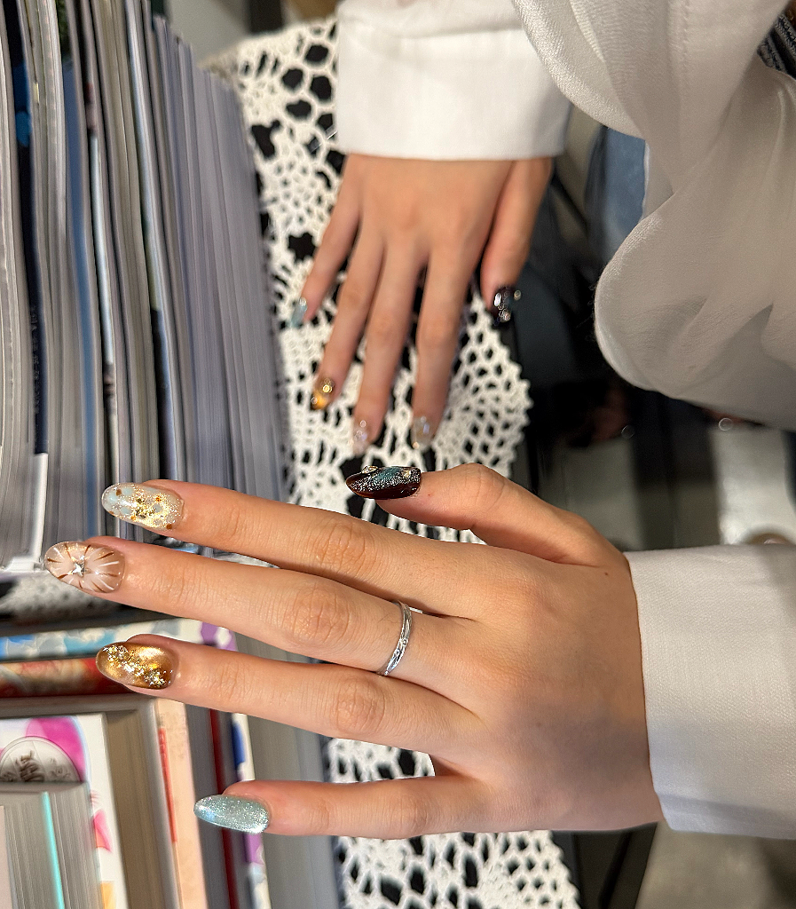 A close-up of a hand with decorative press-on nails, featuring a light blue glittery nail, a gold glittery nail, a clear rhinestone nail, and a dark metallic glittery nail. The person is wearing a white long-sleeve shirt and a silver band ring.
