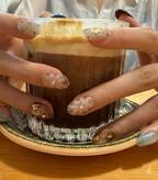 A close-up of hands with decorated almond-shaped nails holding a glass of layered coffee or espresso with foam on a saucer.